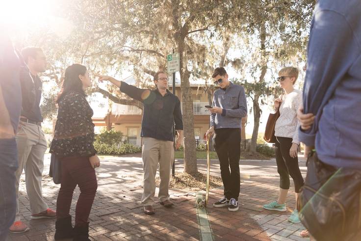 Professor Dan Myers standing on a brick sidewalk pointing to a point of interest while leading students on a digital tour. 