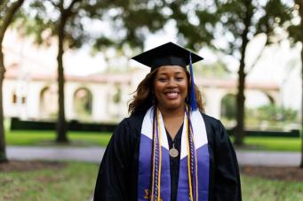 Student in cap and gown standing on campus, smiling at camera.