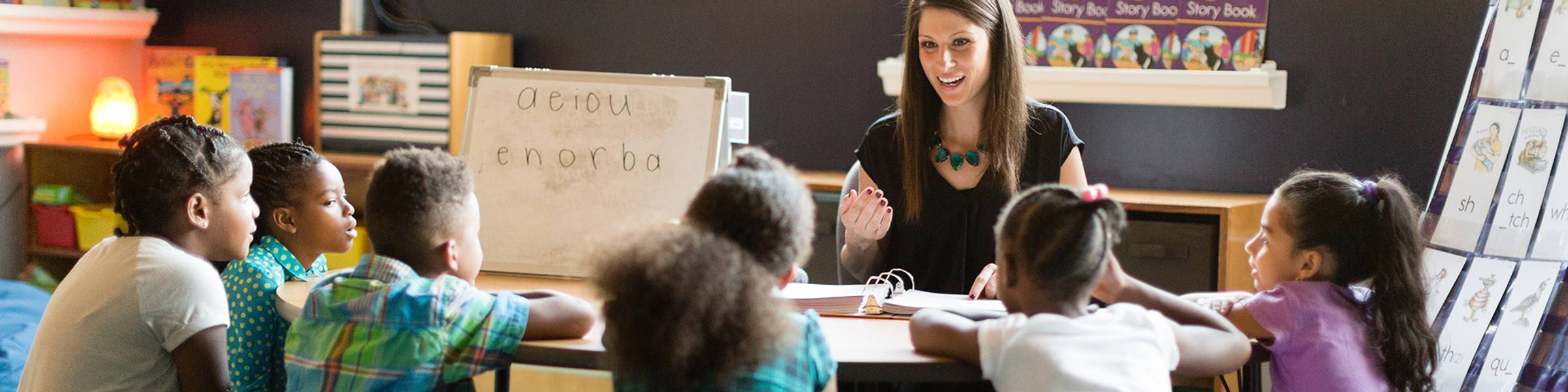 Rollins student sits at a table with six elementary students, teaching the alphabet.