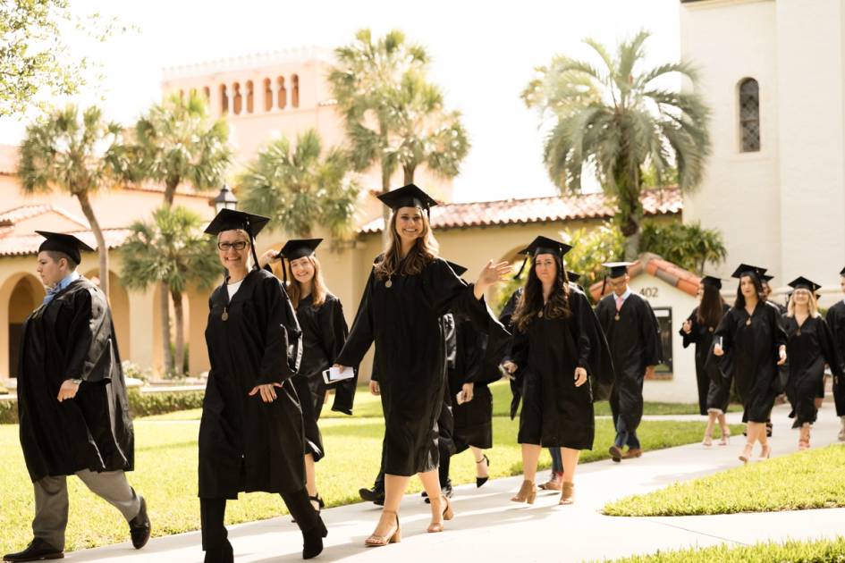 A line of college graduates walk toward a commencement ceremony wearing caps and gowns.