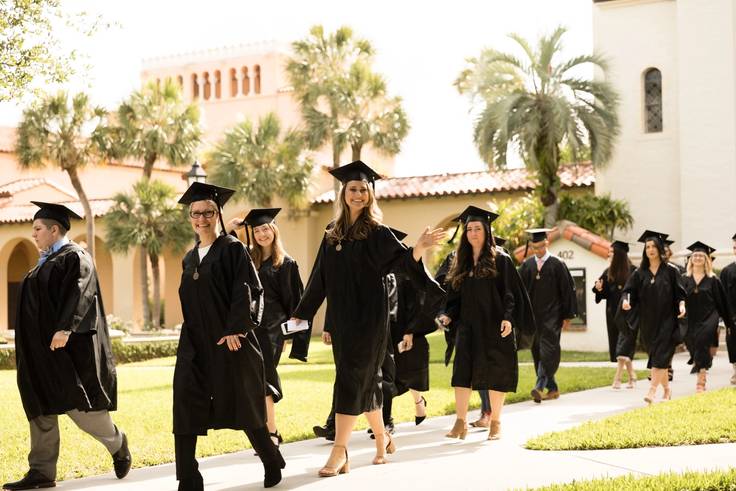 A line of college graduates walk toward a commencement ceremony wearing caps and gowns.