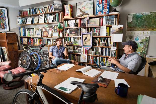 A college professor and graduate student talking in his book and bike filled office.