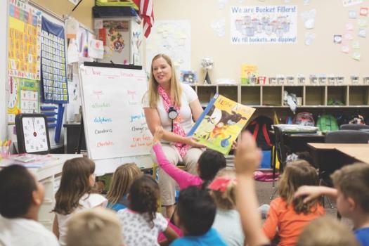 A master of teaching student sits in the center of the classroom reading to a small group of younger children.