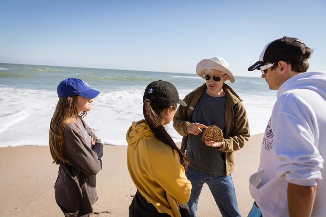 A professor points to a piece of coral that he's holding as he explains a concept to three students.