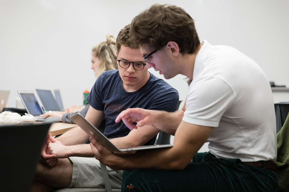 A graduate assistant holding his laptop computer discussing a tiny home design with another student.