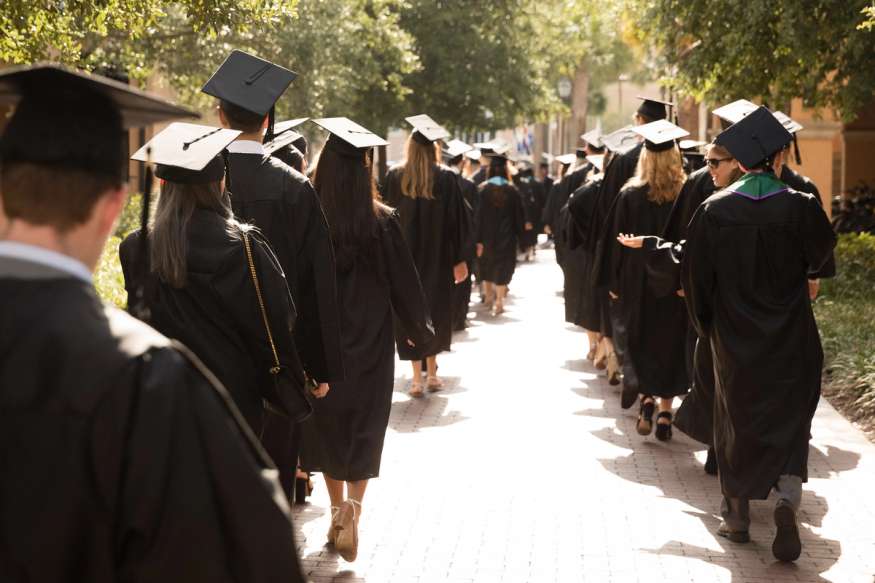 Two lines of college graduates walk to a commencement ceremony wearing caps and gowns.