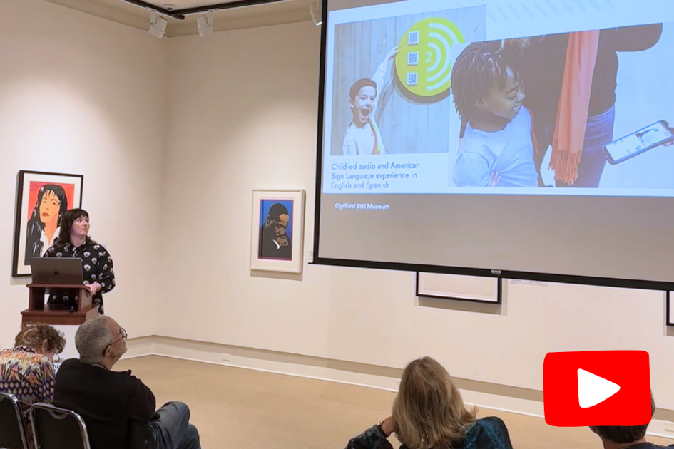 Woman stands at a podium in a art gallery next to a projection screen that features two children and artwork