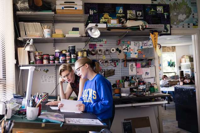 An art history professor working with a student while seated at a studio desk.