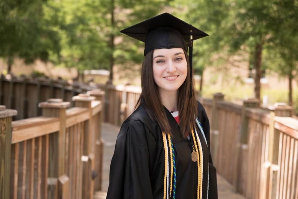 A female college student poses in a cap and gown on campus.