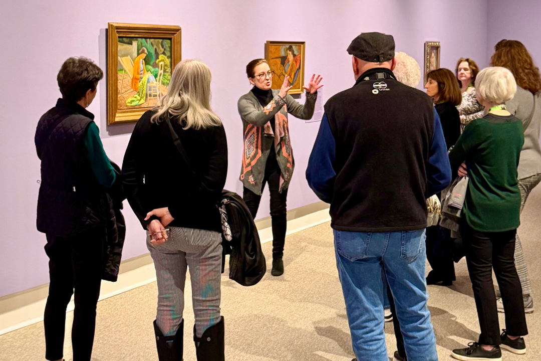 Thuvia Martin raises her arms while giving  tour at Rollins Museum of Art to a group of people. Purple walls with paintings
