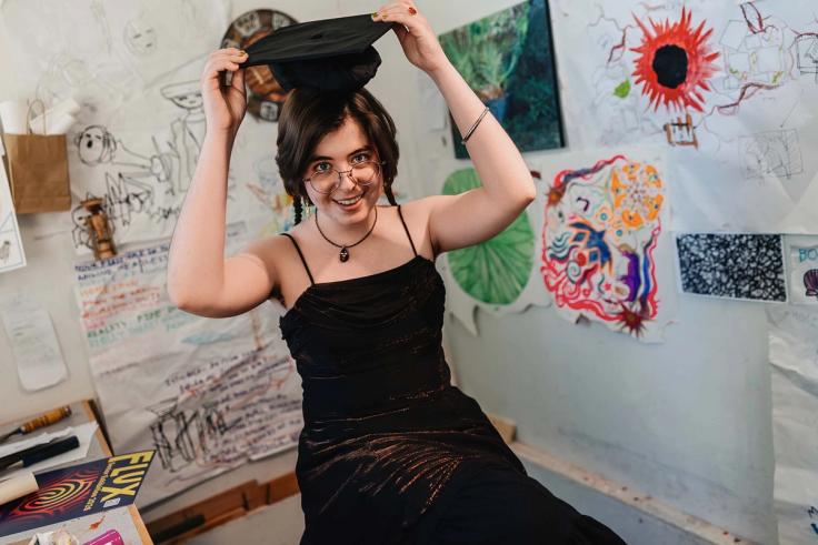 Julia St. John playfully puts on a graduation cap in an art-filled room.