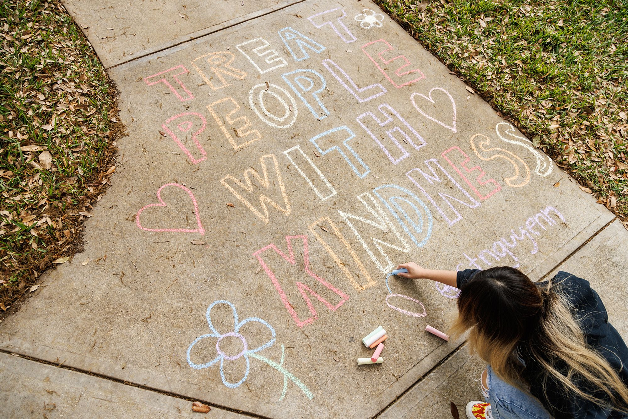 Student writing "Treat People With Kindness" on sidewalk with chalk