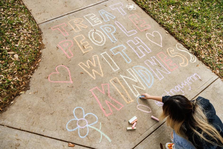 Student writing "Treat People With Kindness" on sidewalk with chalk