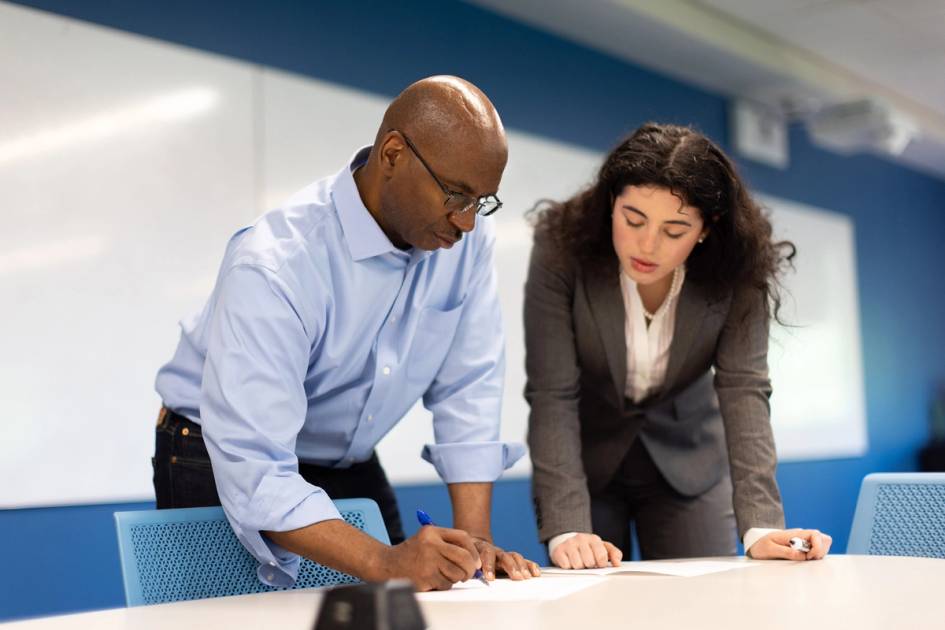 A professor and a student reviewing documents together in a classroom.