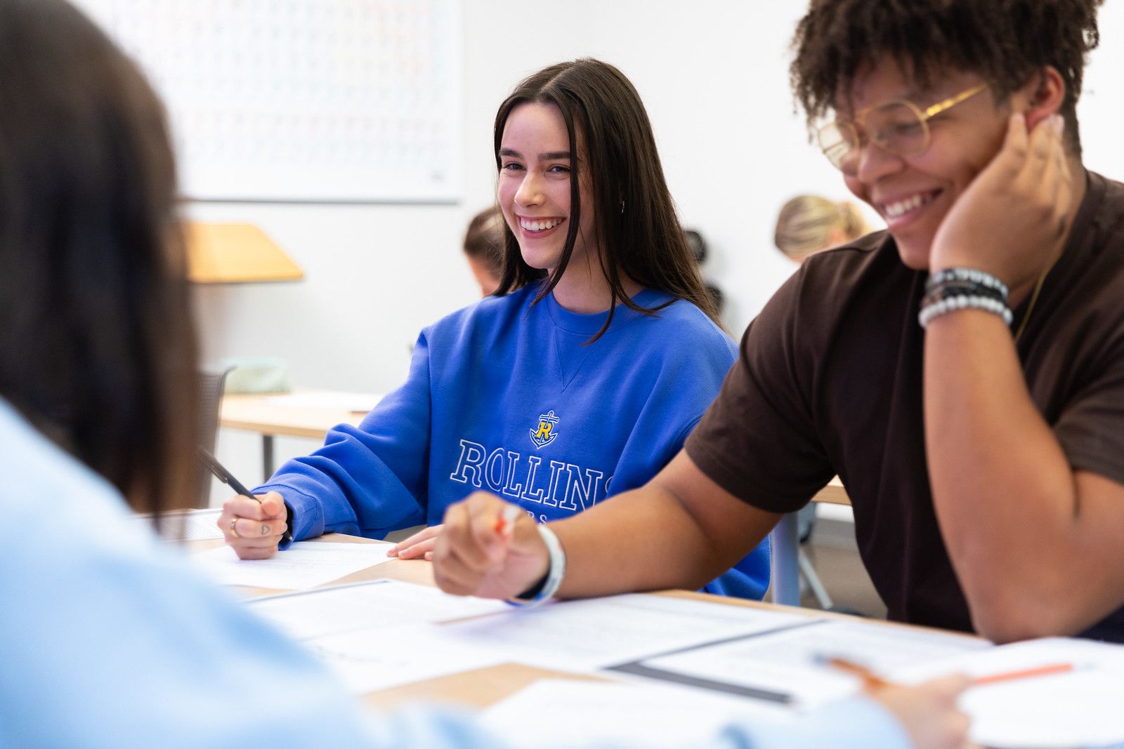 Students collaborating in class, taking notes and laughing. The student in focus wears a royal blue Rollins hoodie.