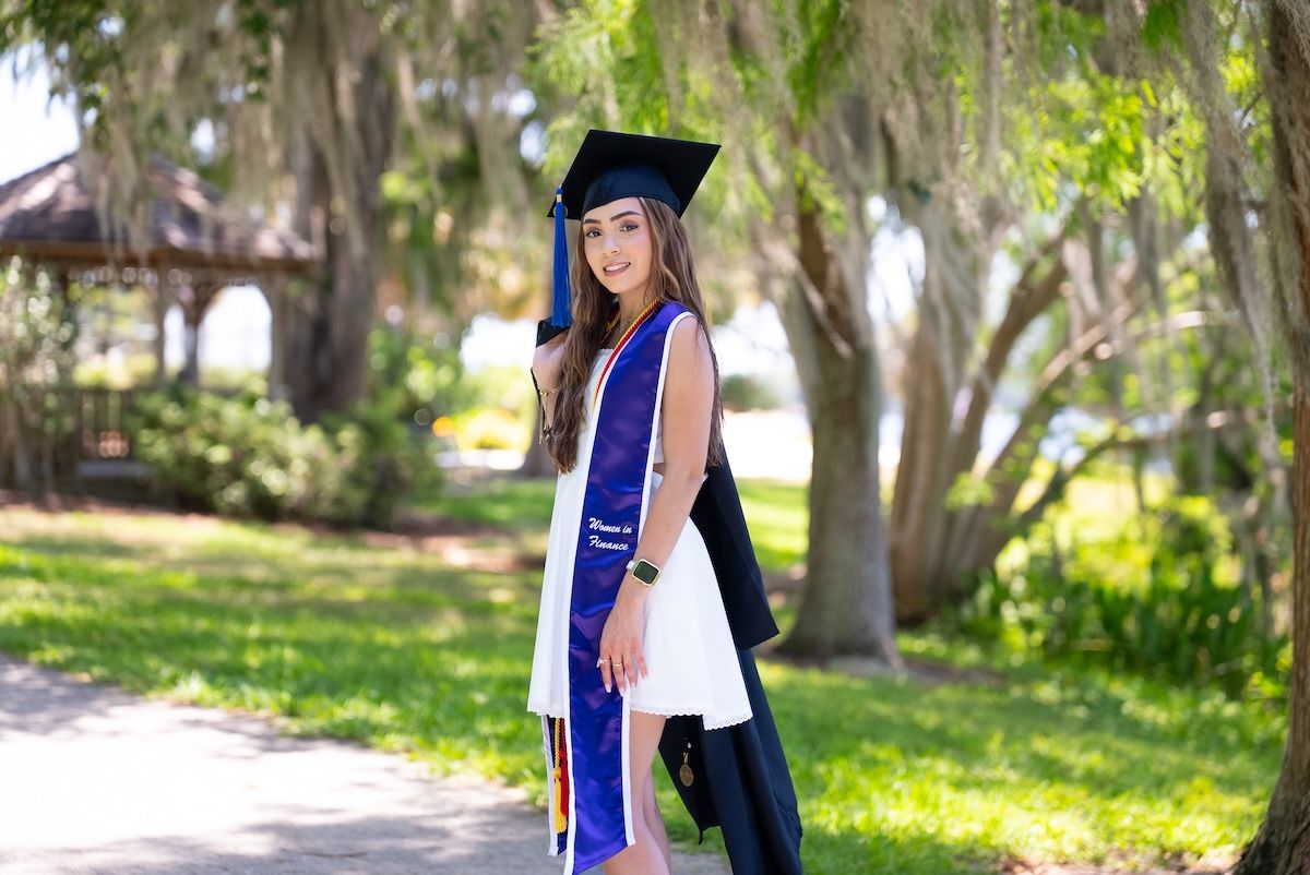 Saarah Valentin, a recent Rollins grad, wearing cap, gown, and honors stole poses outdoors.