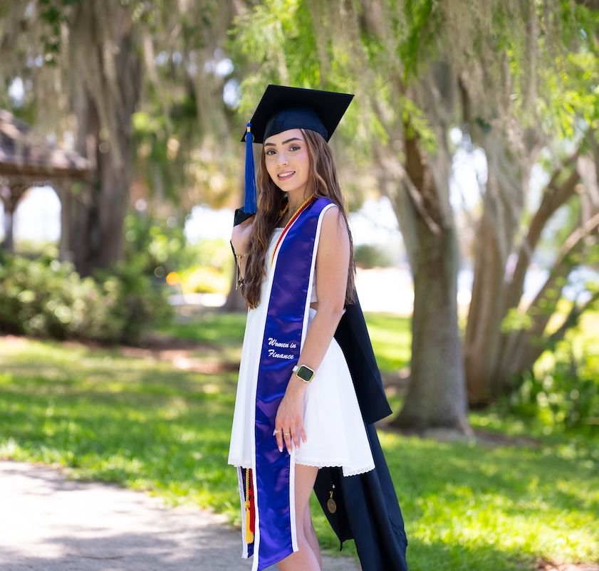 Saarah Valentin, a recent Rollins grad, wearing cap, gown, and honors stole poses outdoors.