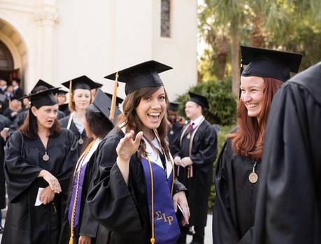 A college graduate in cap and gown waves to the camera before a commencement ceremony.