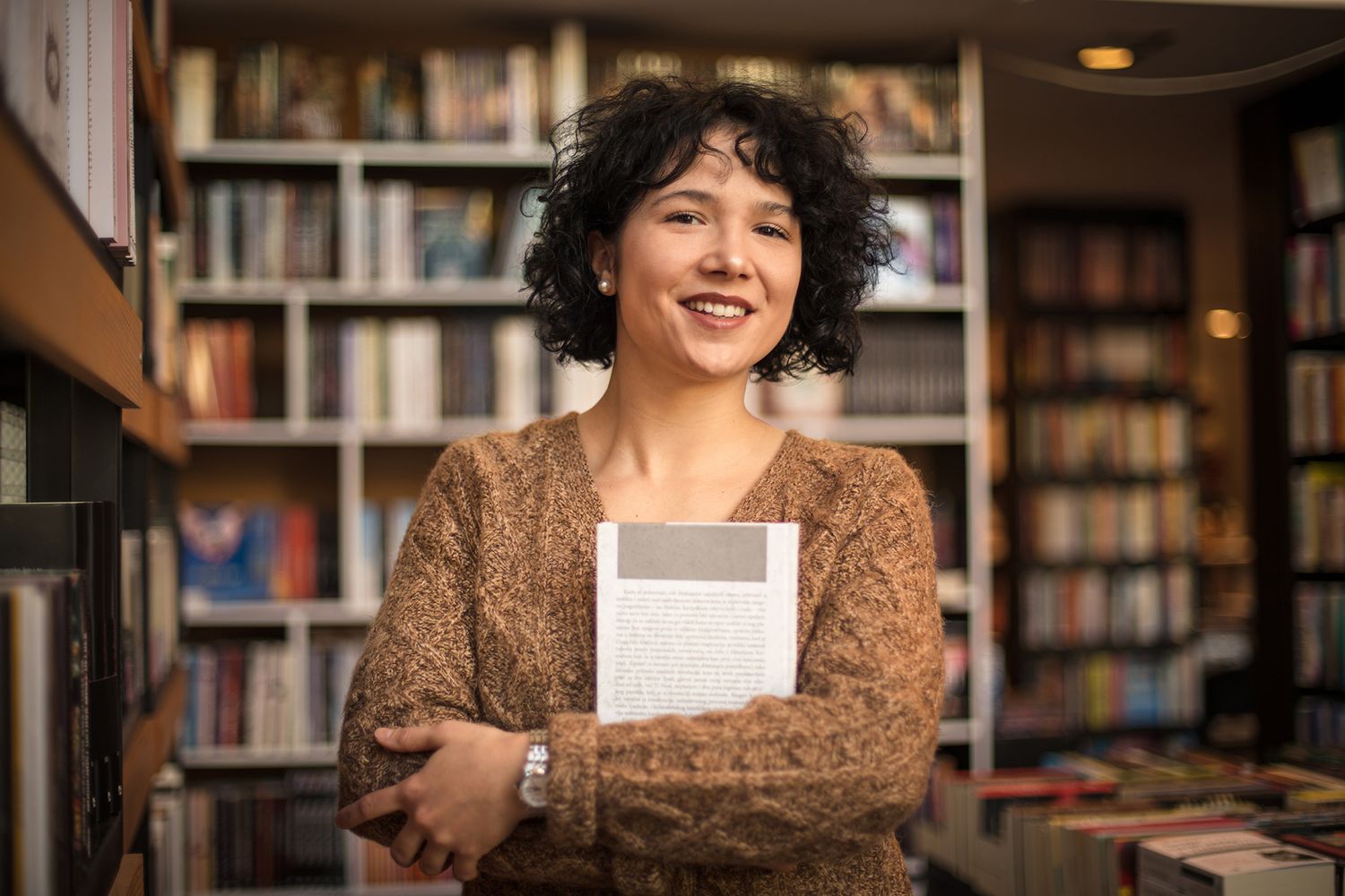 A librarian holds a book across her chest as she stands in front of a shelf of books.