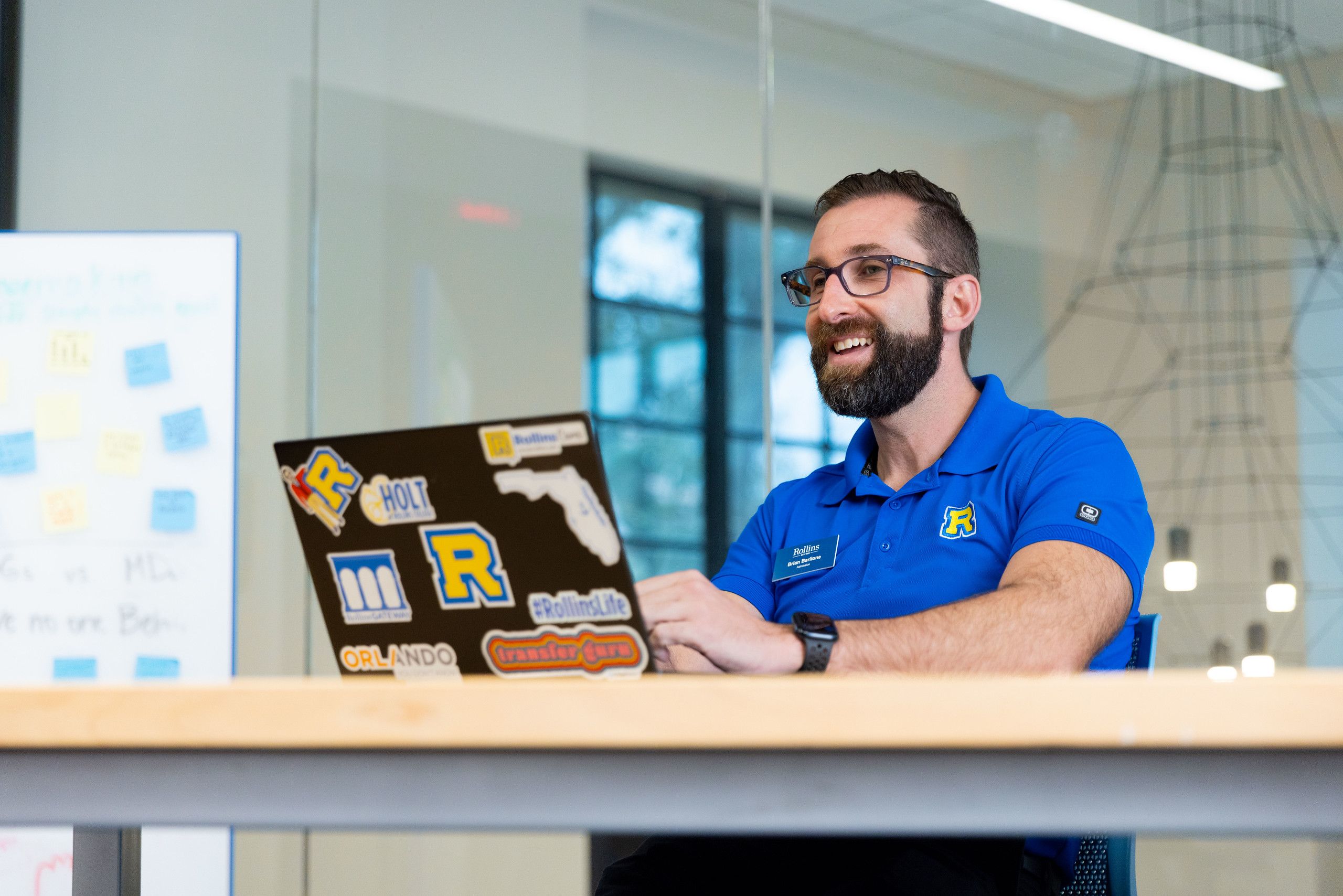 A Rollins counselor in a blue polo shirt sits at a desk, smiling at his laptop