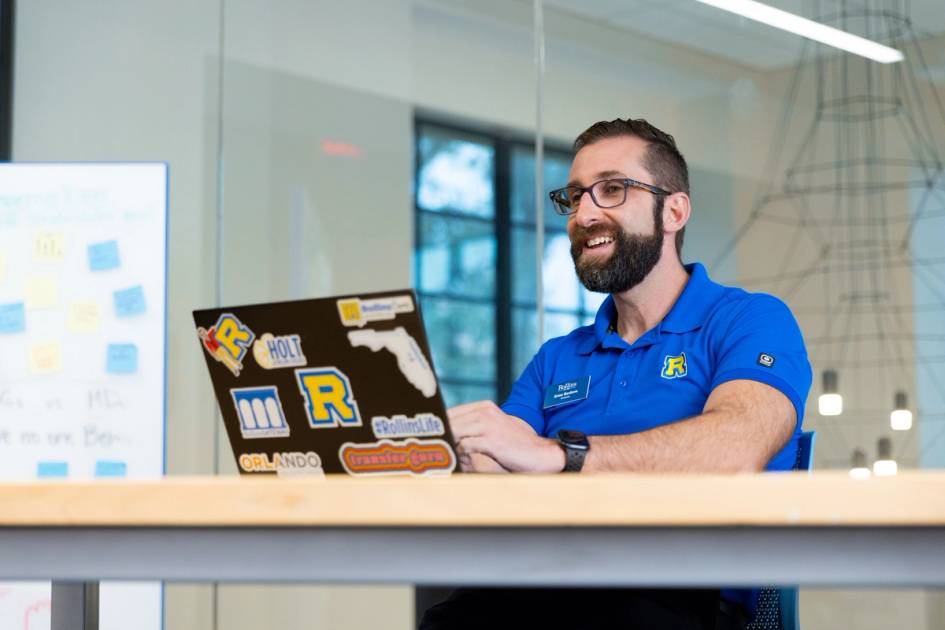 A Rollins counselor in a blue polo shirt sits at a desk, smiling at his laptop