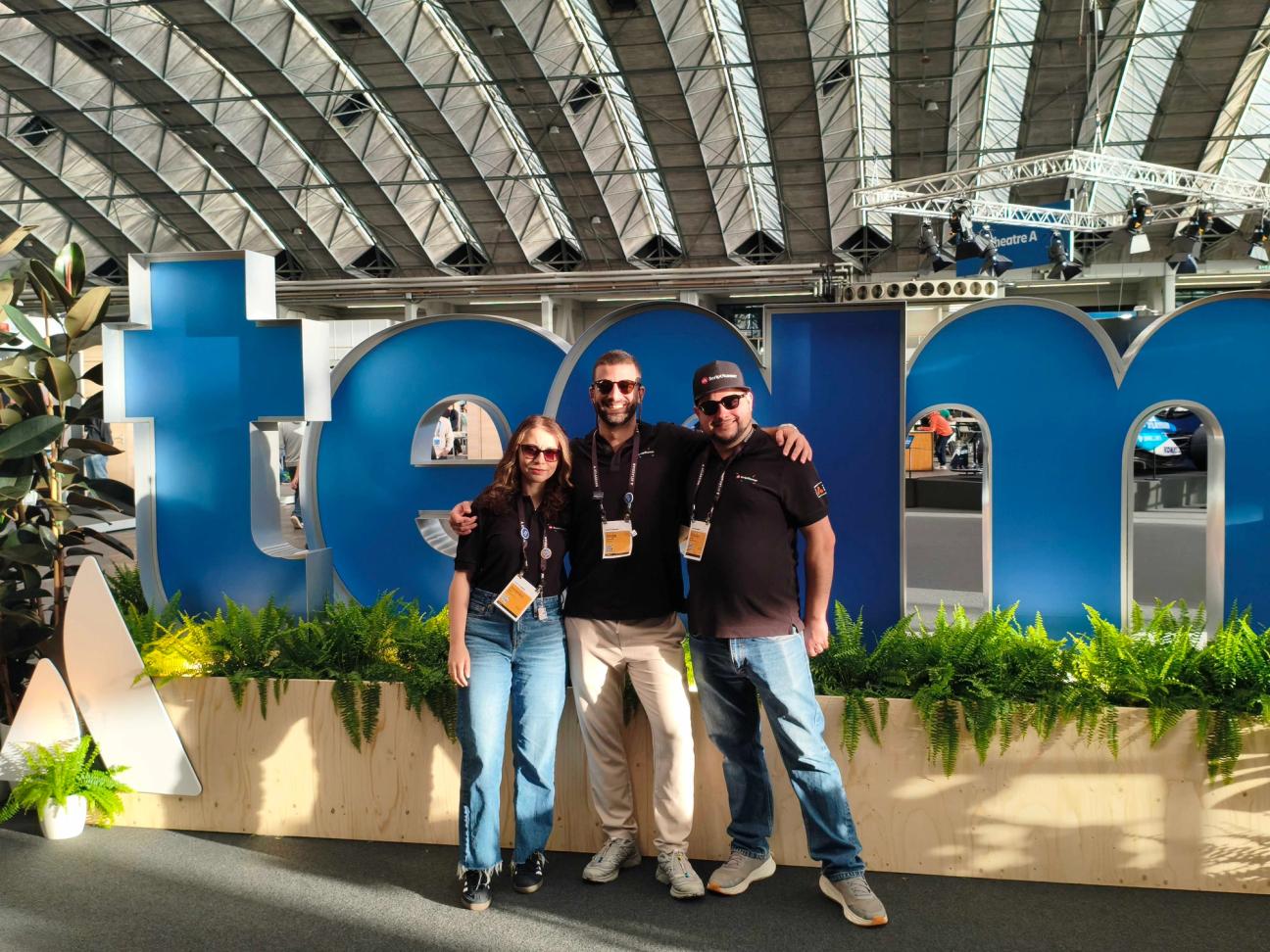 Three people standing and smiling at the team '26 event with a 'team' sign behind them