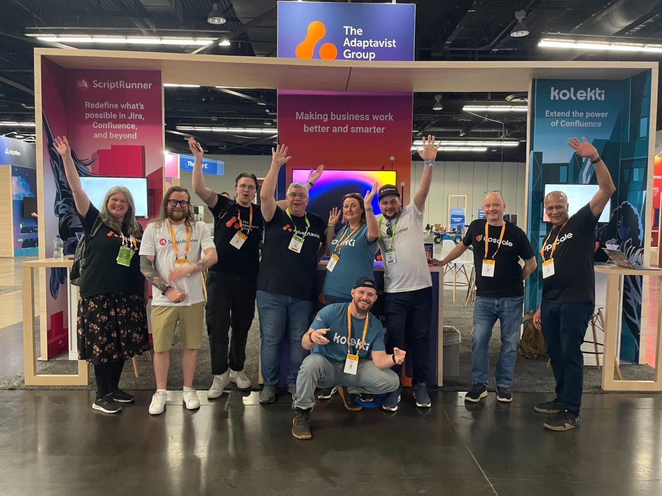 A group of ten smiling people pose enthusiastically with raised arms in front of a booth for The Adaptavist Group at a convention. The booth features branding for ScriptRunner, Kolekti, and other tools. They are at Atlassian Team '25.