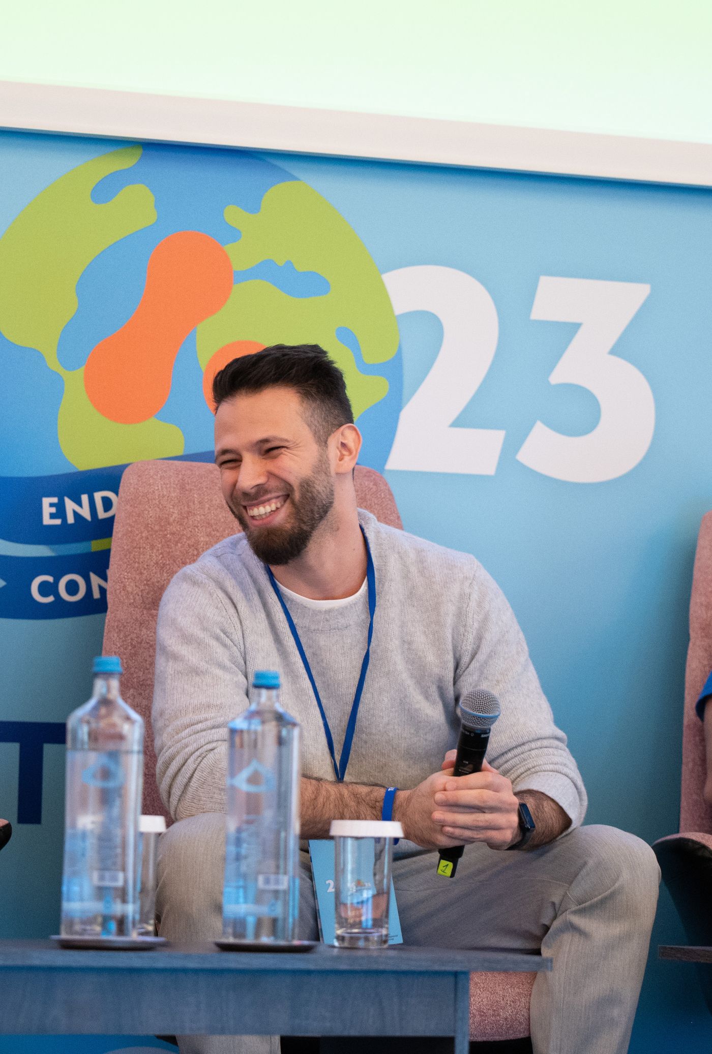 Adil Nasri sitting on a conference stage laughing, holding a microphone