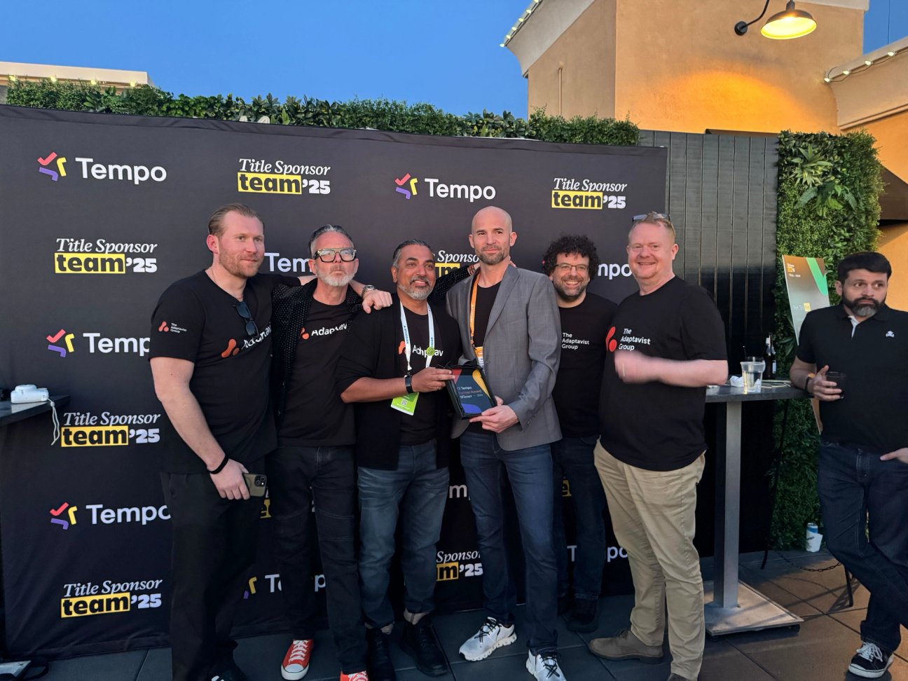 A group of seven TAG colleagues posing together in front of a black backdrop with 'Tempo' and 'Title Sponsor team'25' logos. They are celebrating a win.