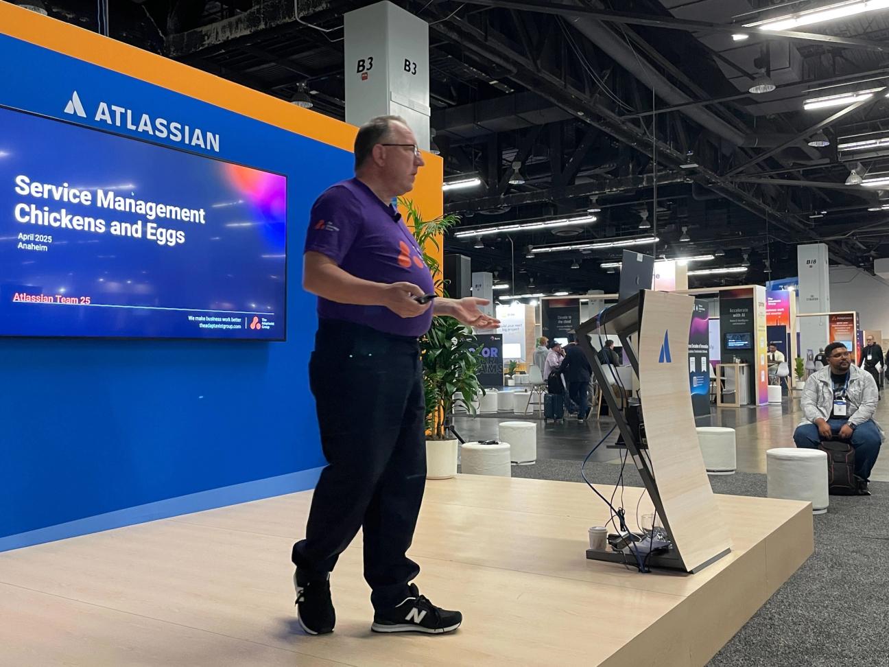 Phil Fox presenting a session titled 'Service Management: Chickens and Eggs' at an Atlassian event, with attendees and exhibition booths in the background.