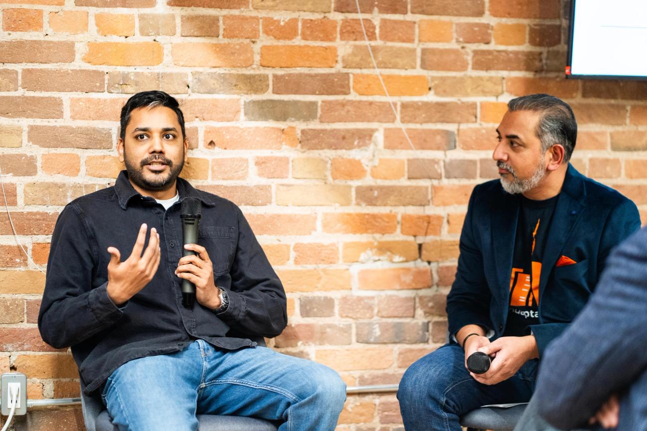 Mayank Agarwal holds a microphone and talks to the audience while sitting next to Harp Athwal