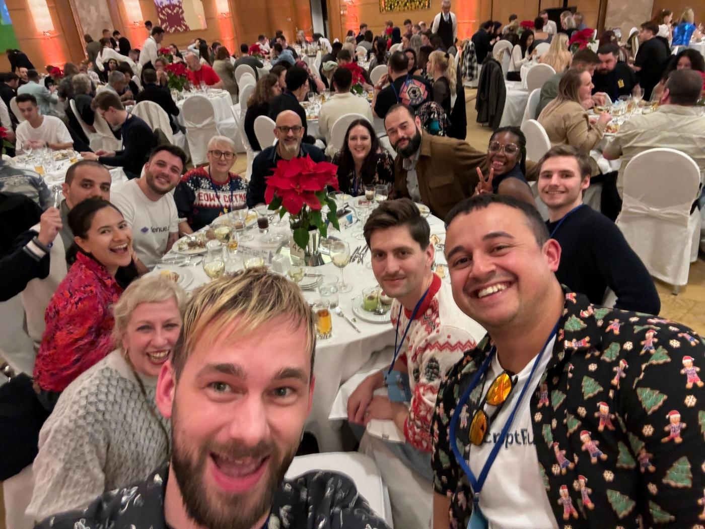 A group of smiling people seated at and standing around a table in a large hall, with lots of other busy tables in the background.