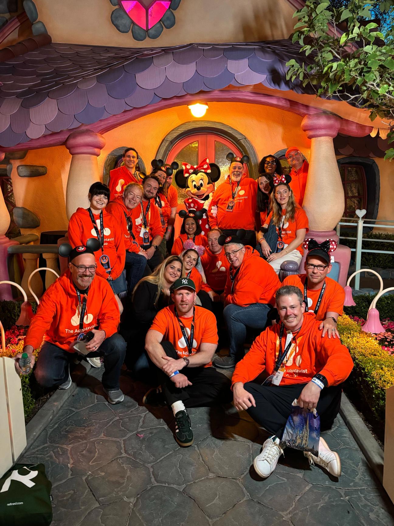 A cheerful group of people posing together in front of Minnie Mouse’s house at Disneyland, with Minnie Mouse herself in the centre. Most of the group are wearing matching bright orange 'The Adaptavist Group' hoodies and some have Mickey or Minnie ears on. The setting is colourful and whimsical, with heart-shaped decorations and vibrant flowers surrounding the house entrance. The group appears happy and excited.