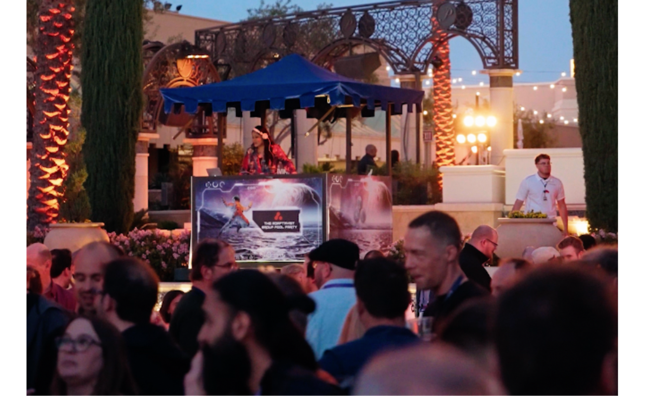 A large crowd of people at an outdoor party, with a DJ booth in the background