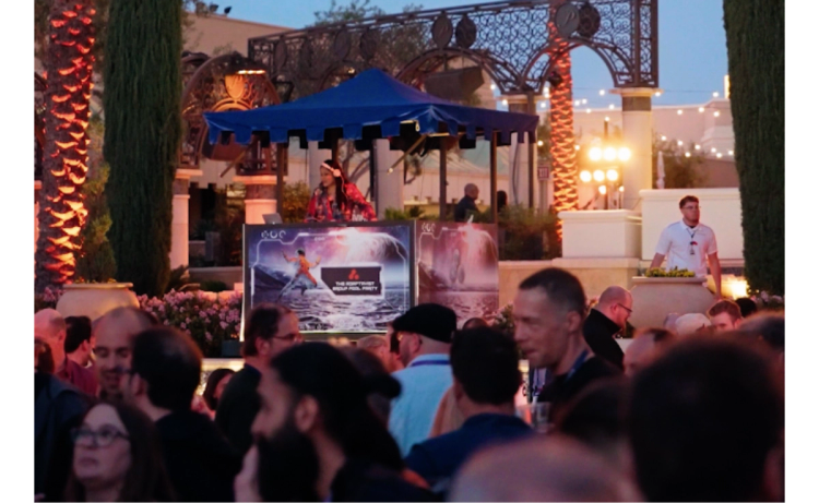 A large crowd of people at an outdoor party, with a DJ booth in the background