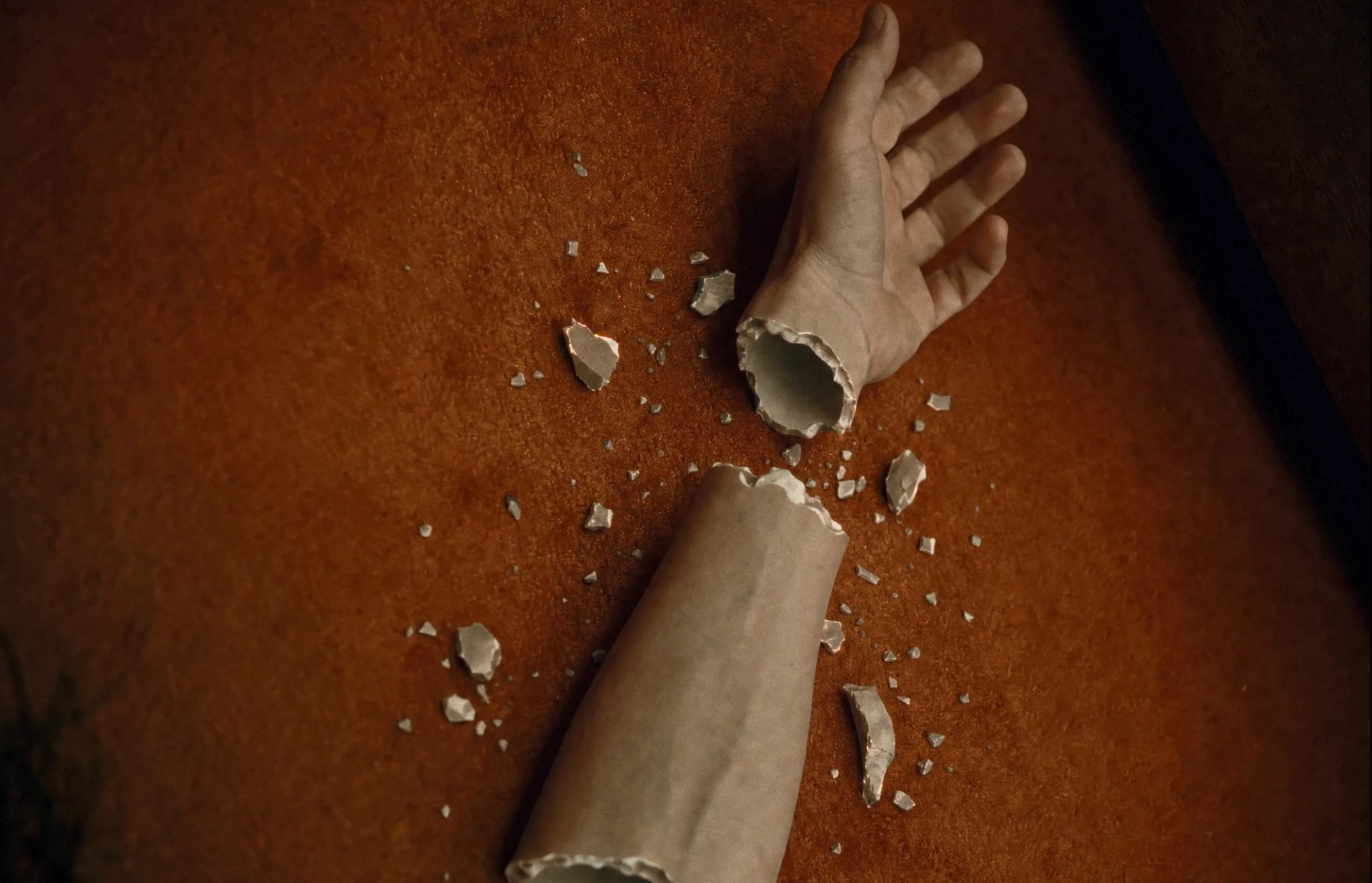 Close-up of a Elisa's hand and forearm broken apart at the wrist, revealing hollow, ceramic-like edges amid shards on an orange carpet.