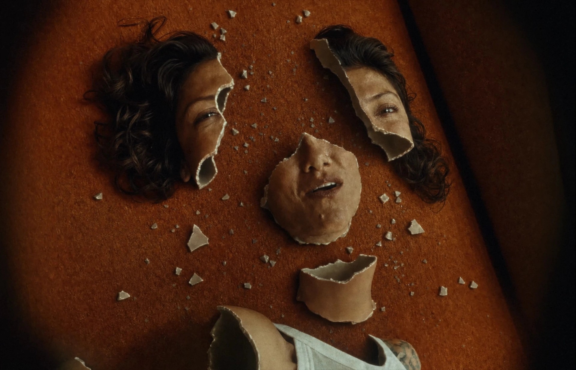 Close-up of a Elisa's face split into hollow, ceramic-like fragments, scattered across an orange carpet among small shards.