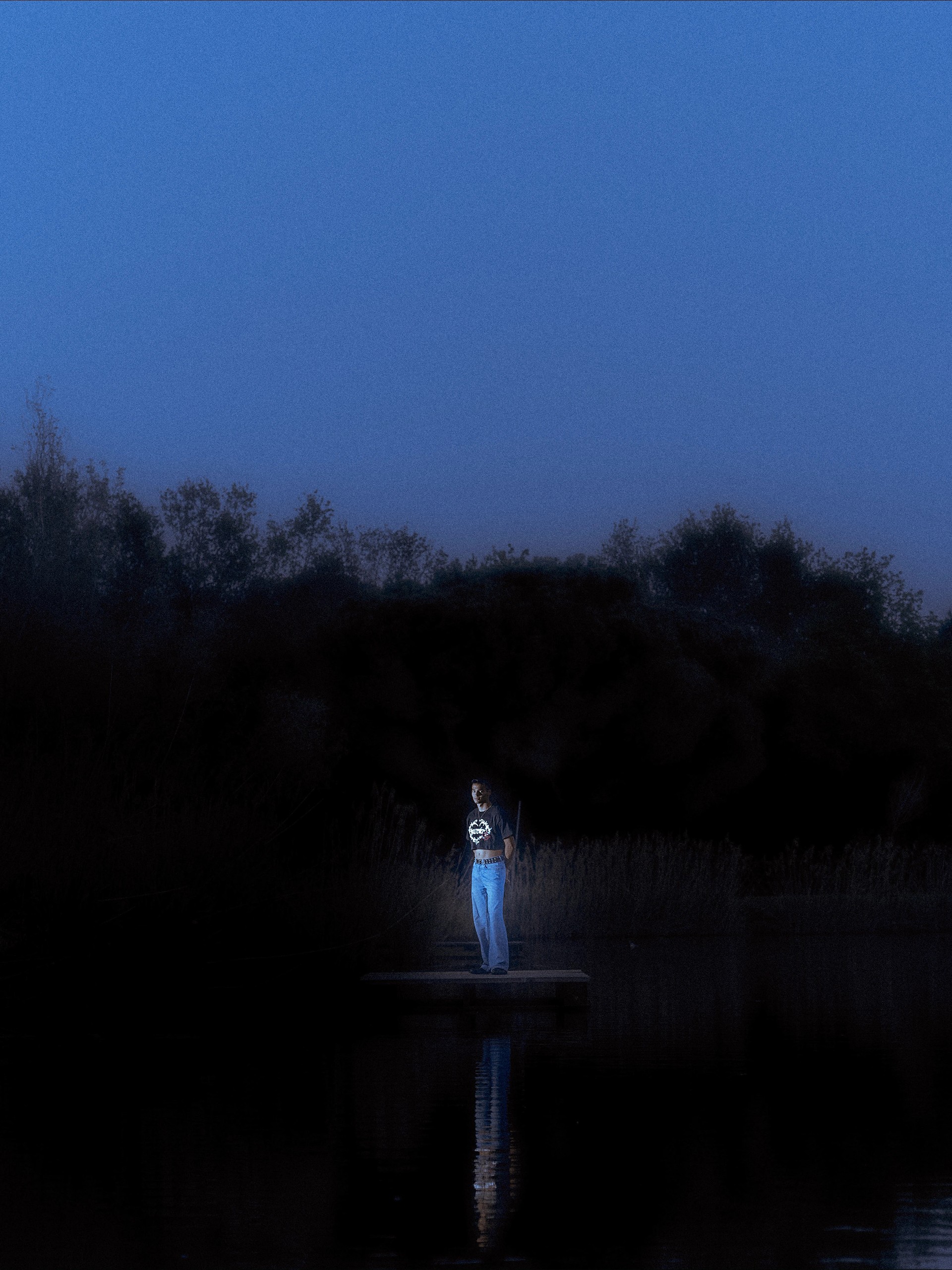Wide dusk shot, the lit figure alone on the jetty above still water