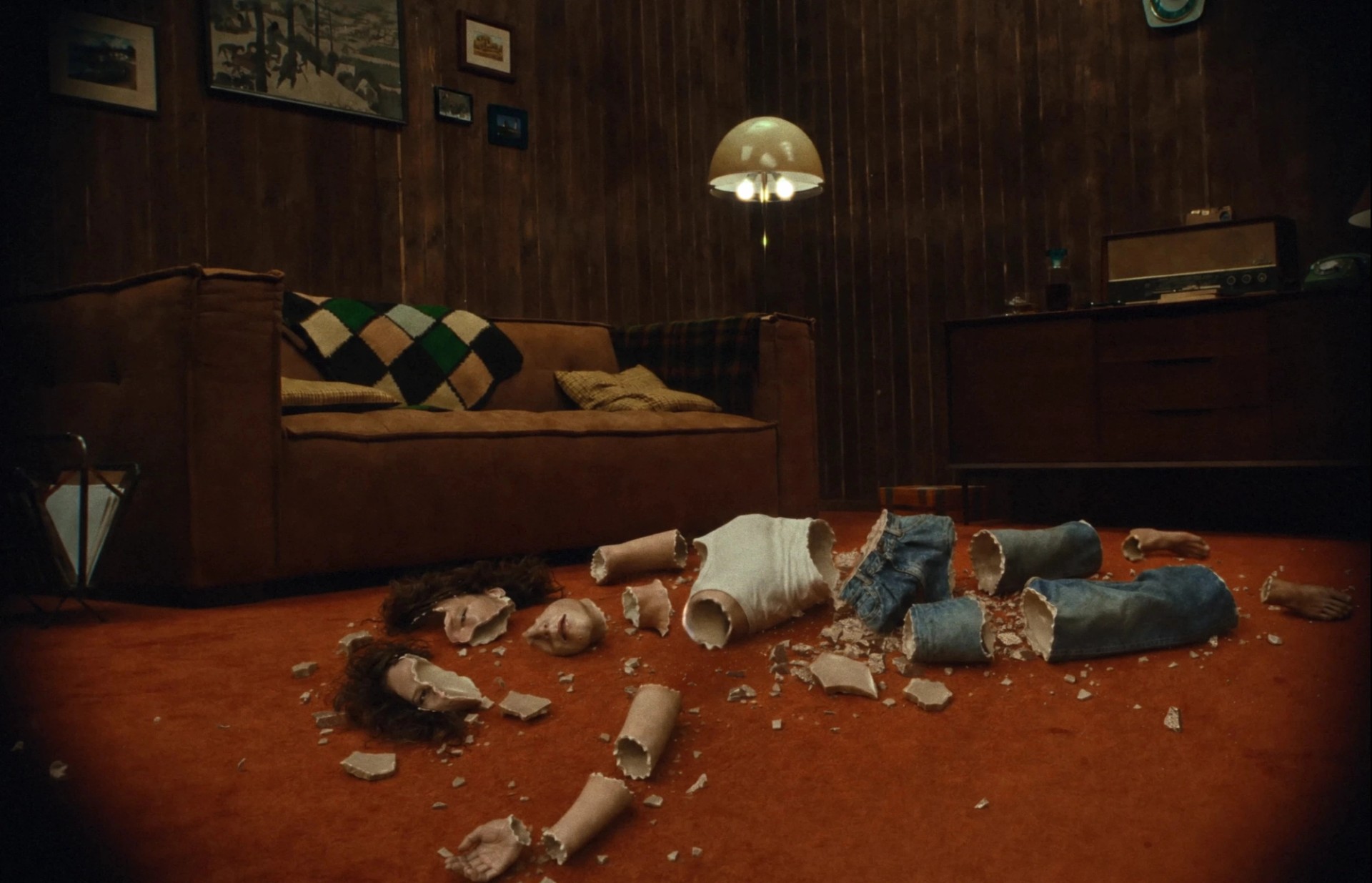 Elisa lies shattered into hollow, ceramic-like pieces on an orange carpet in a wood-paneled living room.