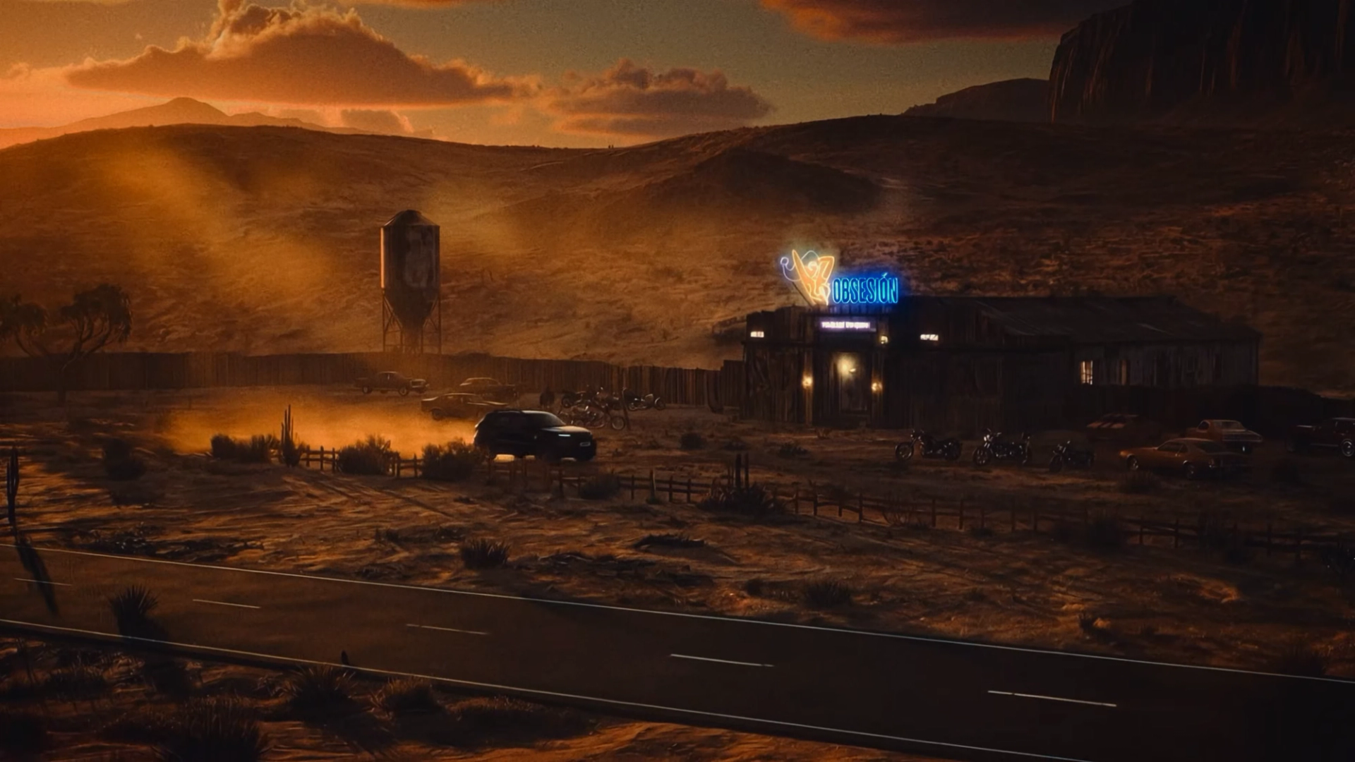 Wide view of an SUV approaching a neon-lit desert bar at sunset, with parked cars, motorcycles, and a water tower.