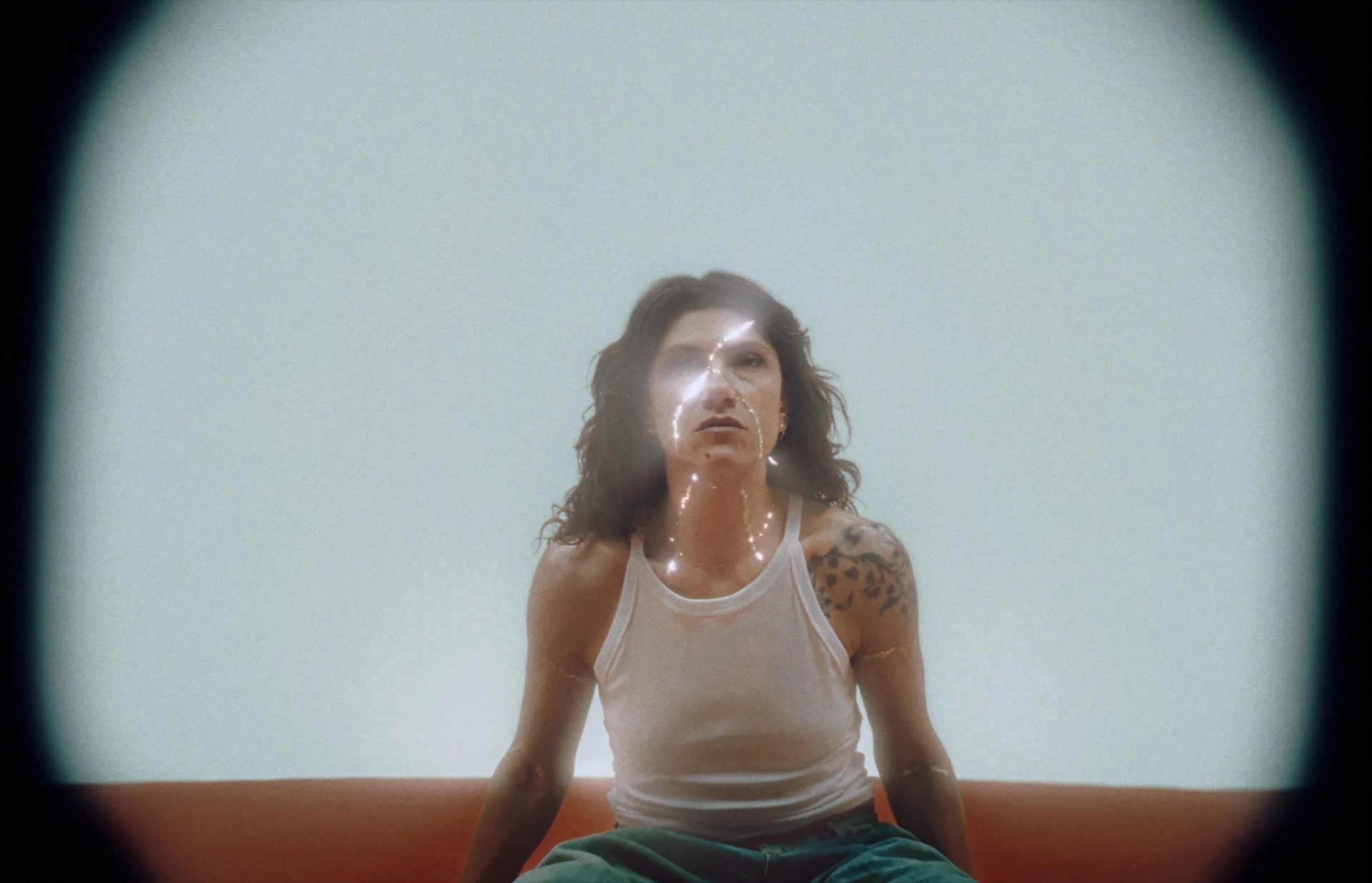 Elisa in a white tank top sits against a pale background, with glowing cracks tracing her face, neck, and arms.