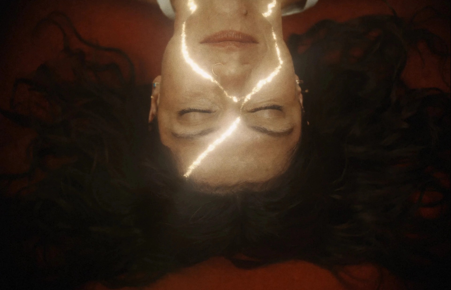 Overhead close-up of Elisa lying on an orange carpet with her eyes closed and glowing golden cracks across her face.