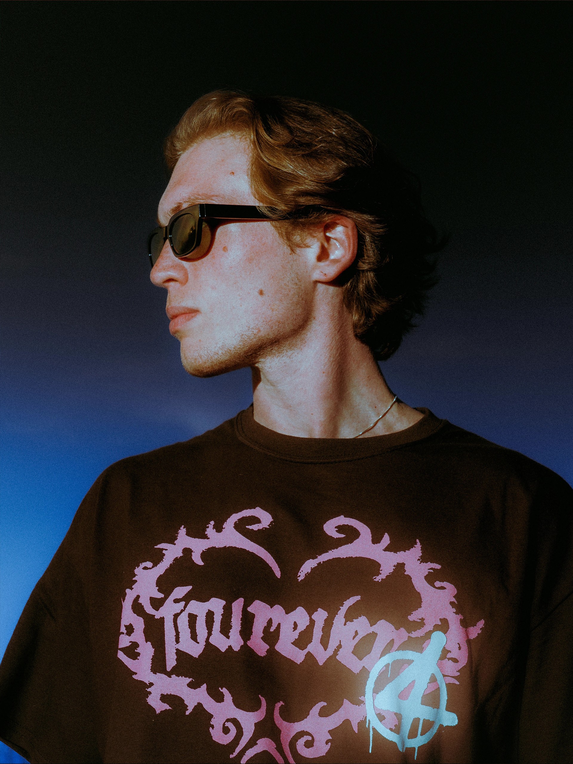 Model in dark sunglasses wearing the black "fourever" tee, lit blue at night Close portrait of a model in the "fourever" graphic tee against blue light Night portrait, black tee with the pink "fourever" chest graphic