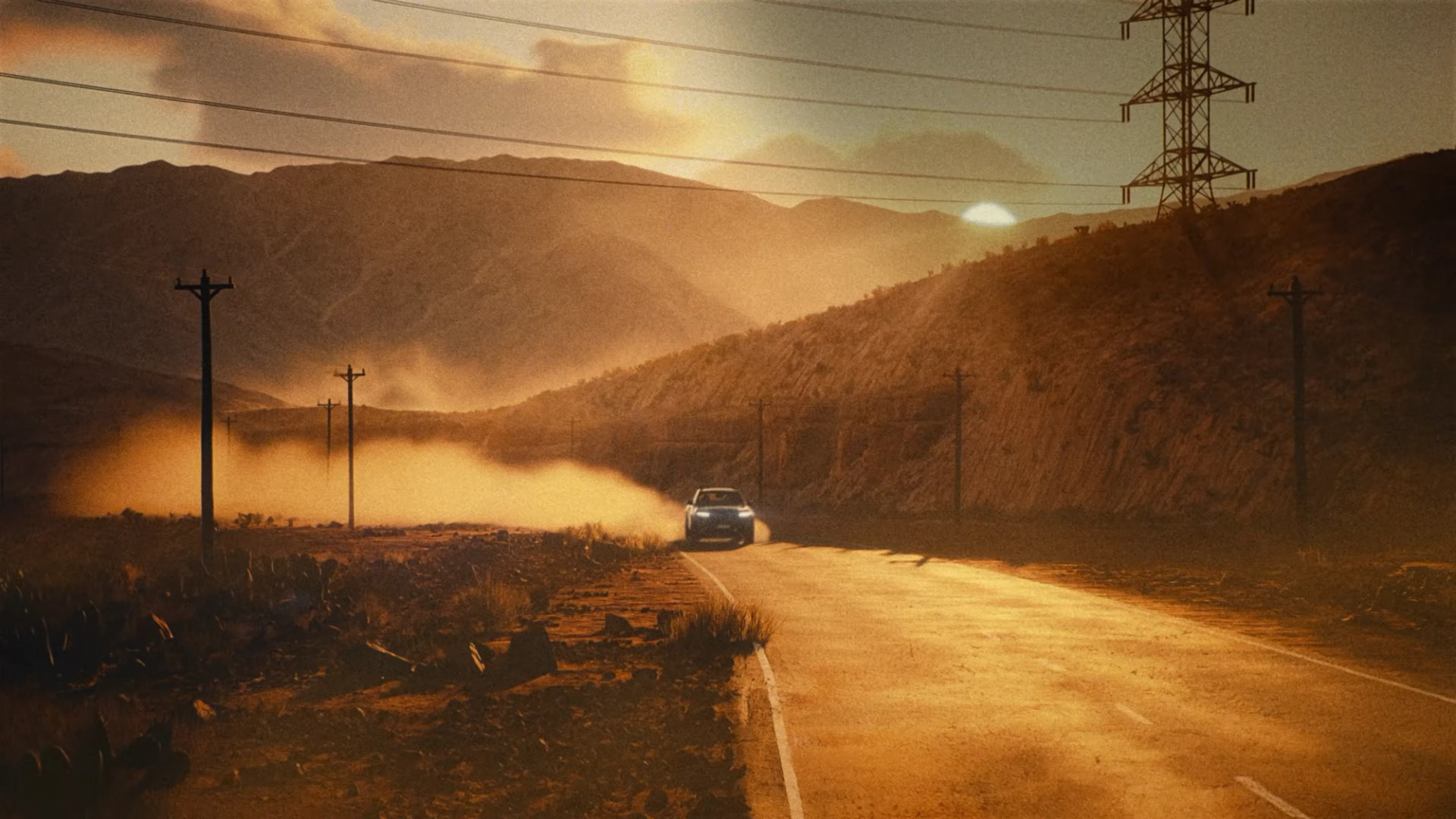 A car drives along a desert road at sunset, trailing a cloud of dust beneath power lines and rugged hills.