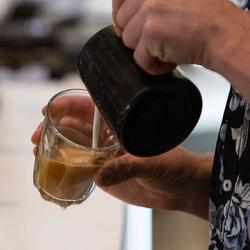 A close-up image of a person pouring hot milk from a small milk jug into a clear latte glass with coffee