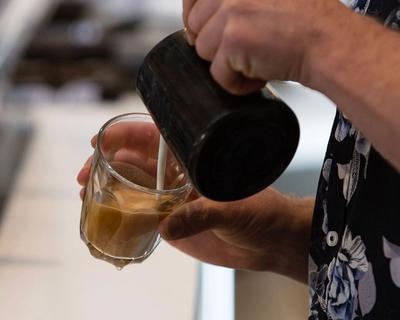 A close-up image of a person pouring hot milk from a small milk jug into a clear latte glass with coffee