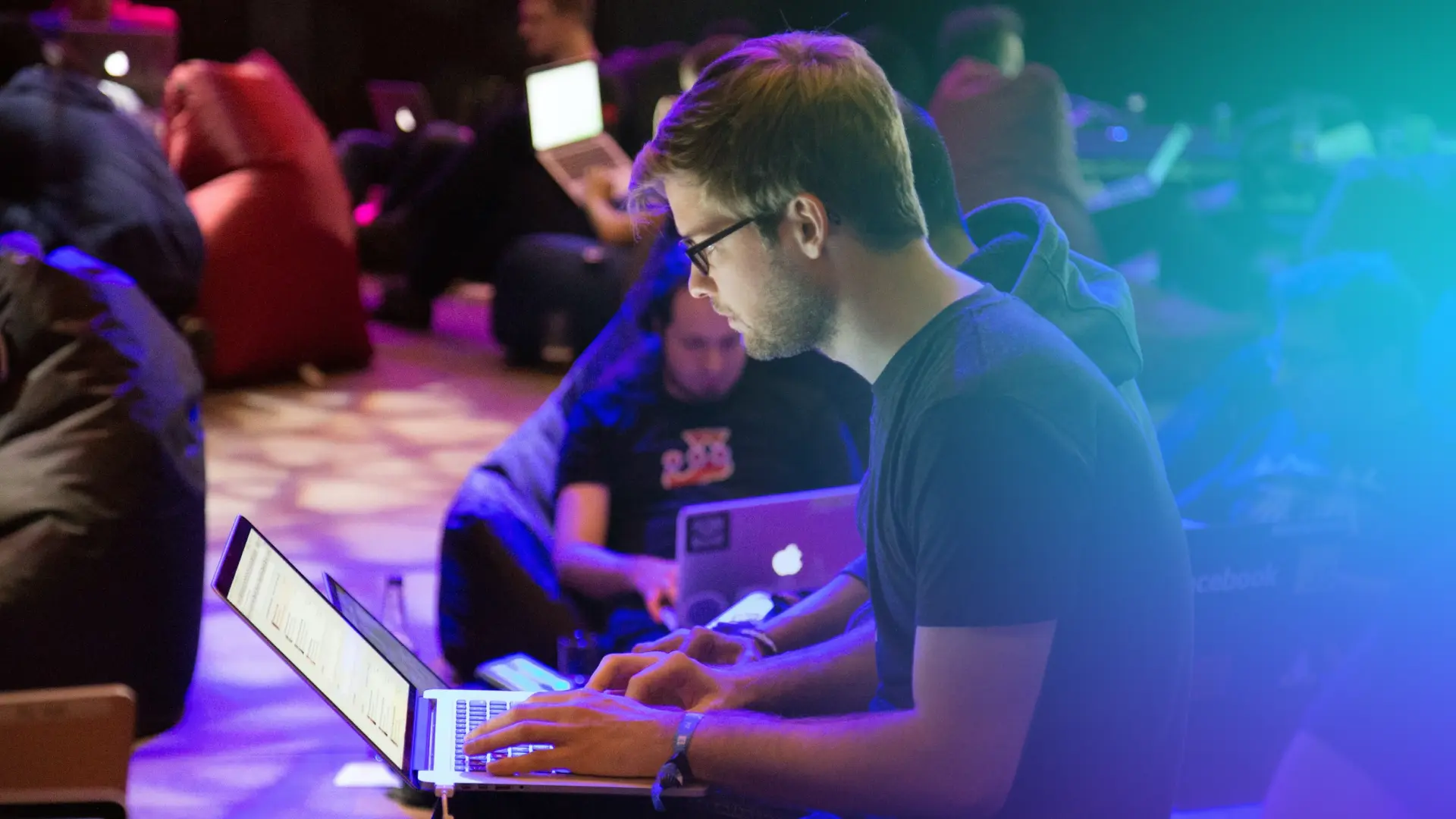 developer with glasses working on a laptop in a packed hackathon room