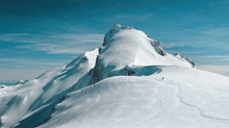 Wide shot of a snowy mountain summit with hikers traversing the ridge against a blue sky.