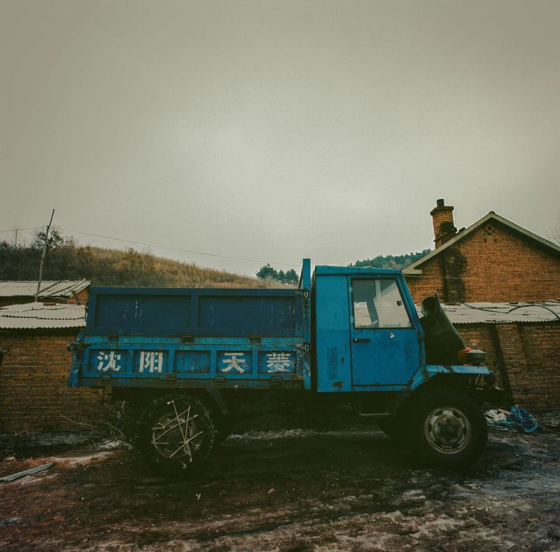 Blue dump truck with Chinese characters parked near brick buildings in a rural landscape.