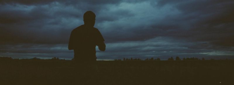 Silhouette of a person standing in a field against a dark, cloudy sky at dusk.